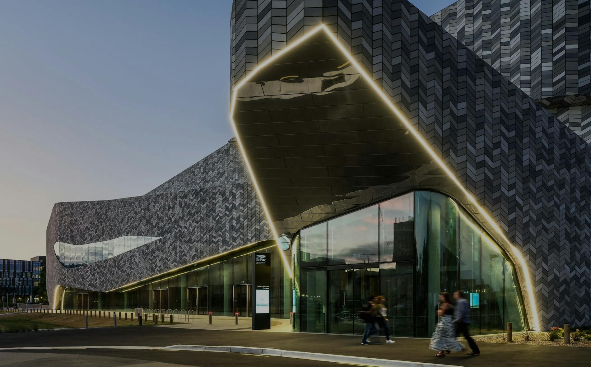 Te Pae, a contemporary building, features a striking, angular facade. People walk past its illuminated entrance, situated in an urban environment during early evening, showcasing modern architecture.